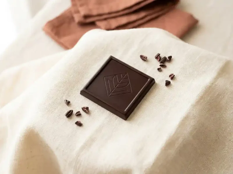Vierkant premium pure chocolade met glanzend gedrukt patroon op crème linnen doek, omgeven door cacaonibs in zacht natuurlijk licht.