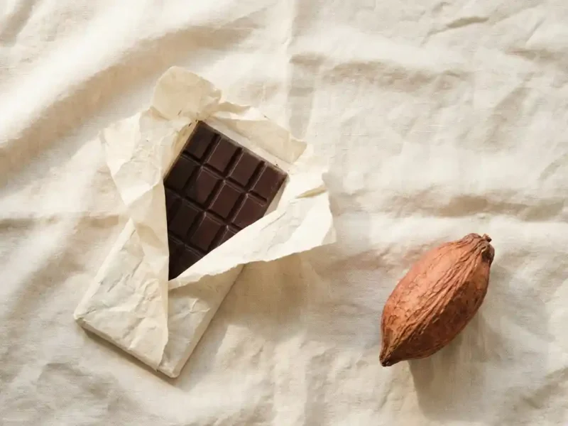 Puur chocoladereep gedeeltelijk uitgevouwen in crèmekleurig papier op linnen ondergrond, met gedroogde cacaopeulen ernaast.